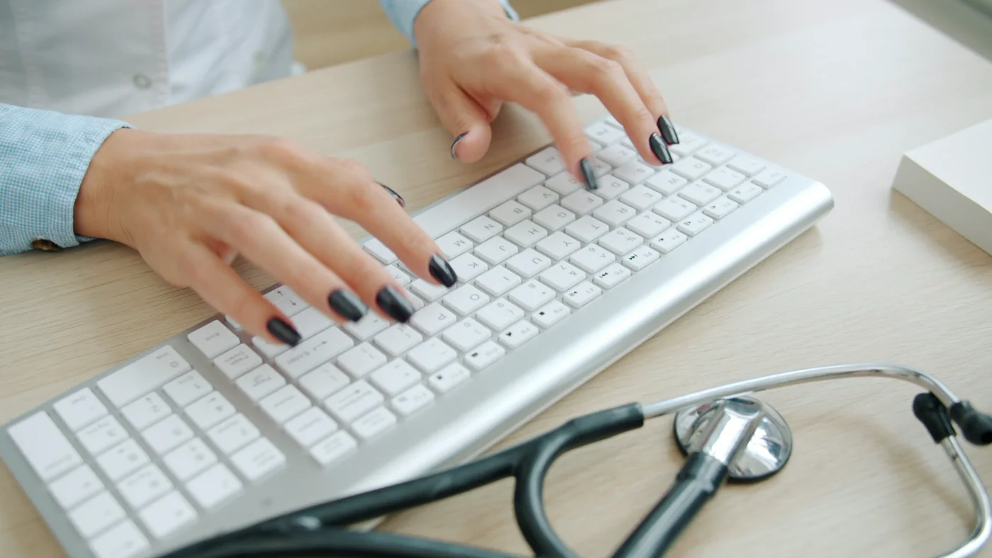 Close-up of hands typing on a keyboard beside a stethoscope